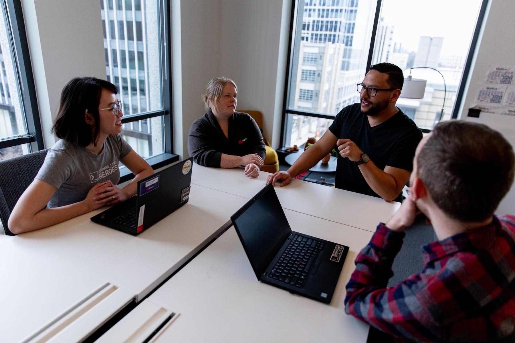 Four people at a desk with laptops having a meeting, listening to someone talk.