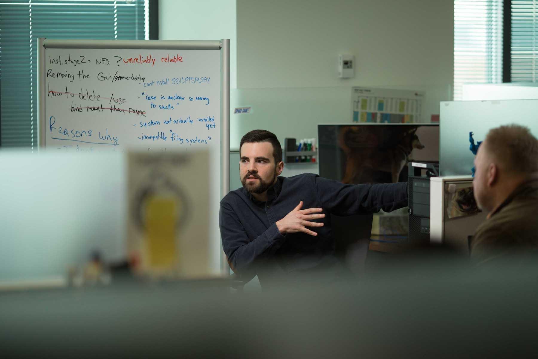 Two people in an office setting in front of a whiteboard, one man is speaking to someone else