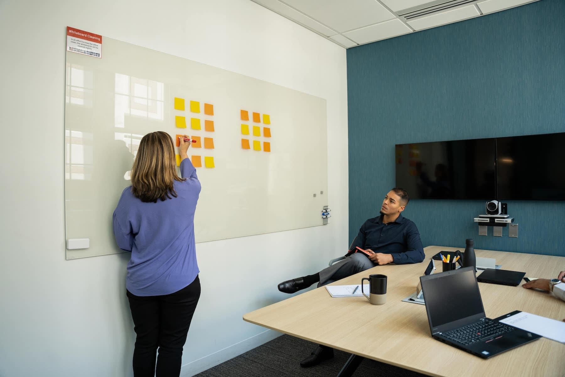 A woman pointing to sticky notes on a whiteboard while a man looks on from a desk